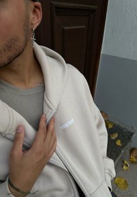 Μπεζ ζακέτα φούτερ με zip, φτιαγμένη από βαμβάκι, με μαλακή υφή, καγκουρό τσέπες και μικρό λευκό λογότυπο στο στήθος. Ορατό casual κάτω ρούχο.