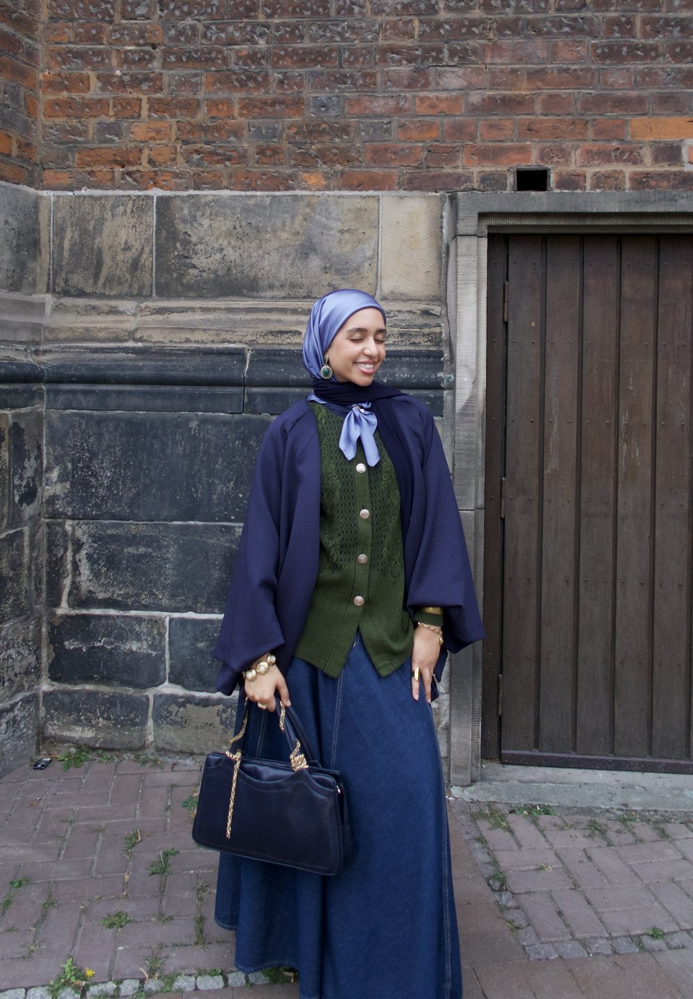 lange denimrok gecombineerd met een groene gebreide cardigan, een marineblauwe cape en een lichtblauwe sjaal. Houdt een donkere handtas vast. Bakstenen muur op de achtergrond.