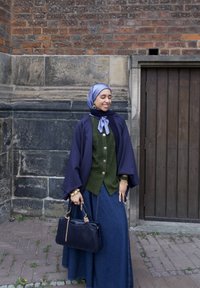 lange denimrok gecombineerd met een groene gebreide cardigan, een marineblauwe cape en een lichtblauwe sjaal. Houdt een donkere handtas vast. Bakstenen muur op de achtergrond.