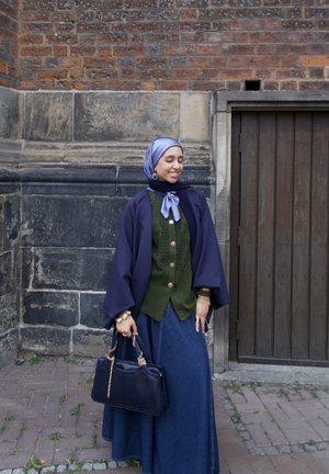 Larga falda de mezclilla combinada con un cárdigan de punto verde, una capa navy y una bufanda azul clara. Lleva un bolso oscuro. Fondo de pared de ladrillo.