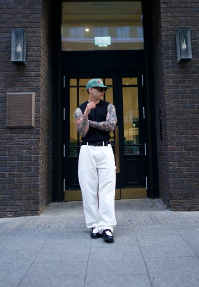 Top negro sin mangas, pantalones anchos blancos, gorra verde y gafas de sol negras; tatuajes visibles en los brazos; de pie fuera de un edificio con paredes de ladrillo.