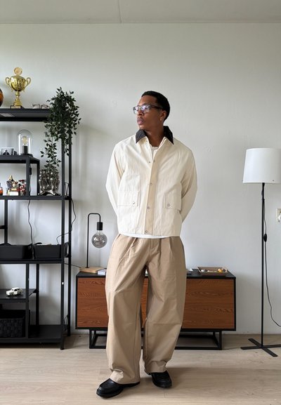 Hombre con gafas, chaqueta beige y pantalones anchos beige de pie con las manos detrás de la espalda en una habitación minimalista con estanterías y lámpara de pie.
