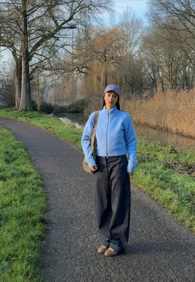 Suéter azul claro esponjoso con cremallera, pantalones anchos negros, zapatos color canela y gorra gris. De pie en un camino con césped y árboles en el fondo.
