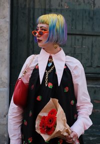 Robe noire avec broderie de fraises, chemise à col rose, cheveux colorés, lunettes de soleil rouges, sac à main rouge et un bouquet de fleurs rouges.