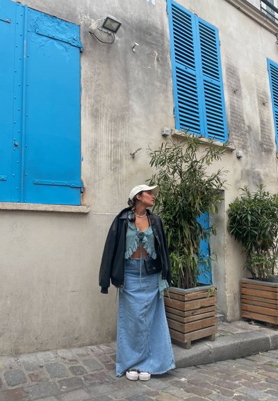 Chaqueta negra oversize, top corto azul claro con volantes, falda de mezclilla larga, zapatillas blancas; frente a una pared texturizada y contraventanas azules.