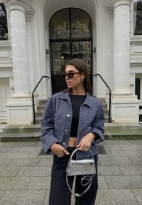 Námořnický pruhovaný sako na knoflíky, černý crop top a tmavé kalhoty. Stříbrná crossbody taška. Sluneční brýle s tmavými želvovinovými obruby. Pozadí šedých kamenných schodů.