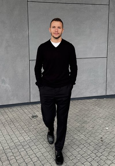 Suéter negro de cuello en V sobre una camisa blanca, combinado con pantalones negros y zapatos negros brillantes; la persona está de pie contra una pared gris.