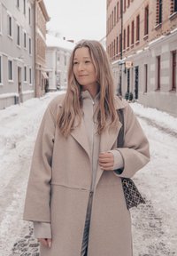 Femme en manteau beige et pull debout dans une rue pavée recouverte de neige, entre des bâtiments en briques et en plâtre.