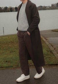 Homme debout près d'un lac portant un long manteau sombre, un pull gris, un pantalon marron, des baskets blanches, les mains dans les poches par une journée nuageuse.