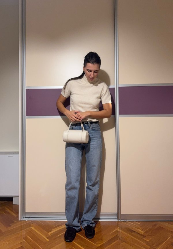 Une femme regarde vers le bas en tenant un petit sac à main blanc. Elle porte un haut crème, un jean bleu et des chaussures noires, à l’intérieur.