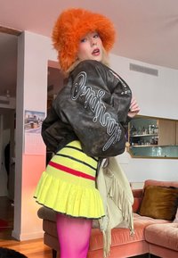 Personne portant un chapeau en peluche orange, une veste bomber foncée avec des inscriptions blanches, une jupe rayée jaune et des collants roses, posant à l'intérieur près d'un canapé.
