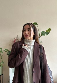 Brun blazer over en creme ribbet turtleneck sweater. Tekstureret stof, afslappet pasform. Brun lædertaske med rem, grønne planter i baggrunden.