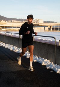 Άνδρας ντυμένος με μαύρα αθλητικά ρούχα και λευκά αθλητικά παπούτσια τρέχει σε ασφαλτοστρωμένο μονοπάτι με χιόνι συσσωρευμένο στο πλάι και κτίρια στο φόντο.