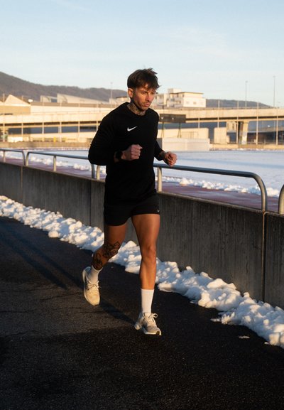 Hombre vestido con ropa deportiva negra y zapatillas blancas trotando por un camino pavimentado con nieve acumulada a los lados y edificios al fondo.