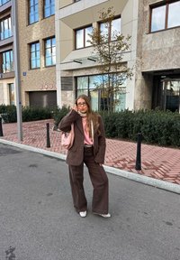 Blazer en velours marron sur un haut rose, associé à un pantalon large marron et des chaussures grises. Tenant un sac rose clair. Arrière-plan urbain.