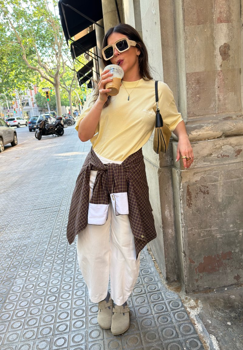 Woman wearing oversized sunglasses and light yellow shirt drinks iced coffee, leaning against stone wall on urban sidewalk.