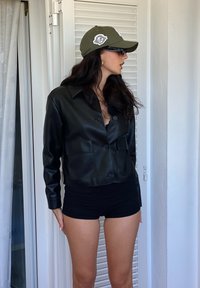 Čierna kožená skrátená bunda s golierom a prednými vreckami, skombinovaná s čiernym priliehavým topom a olivovo-zelenou šiltovkou.