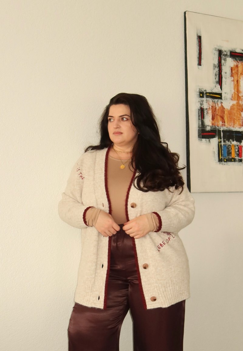 Femme aux longs cheveux foncés portant un cardigan crème, un haut marron et un pantalon bordeaux, debout à l'intérieur près d'une œuvre d'art murale abstraite, regardant sur le côté.