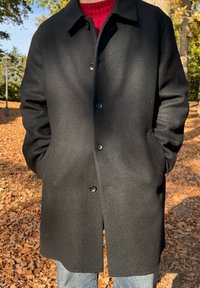 Juodas vilnonis paltas su apykakle, priekinė saga uždarymas ir šoninės kišenės. Dėvimas ant raudono megztinio, fone krentančių lapų.