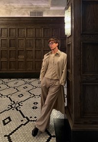 Tan trench coat a béžový cardigan cez svetlé nohavice; čierne topánky; postavený proti drevenej stene v prostredí s vzorovanými dlaždicami.