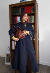 Manteau bleu marine à manches longues avec poches latérales, mettant en valeur la silhouette. Sac à main en cuir marron avec deux poignées. Haut en maille jaune en dessous.