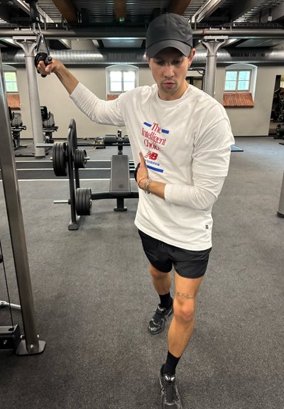 Camisa blanca de manga larga con texto rojo y azul, pantalones cortos negros y zapatillas negras. Persona utilizando equipamiento de gimnasio en una sala de fitness con pesas.