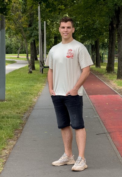 Camiseta de manga corta gris claro con un gráfico rojo, pantalones cortos de mezclilla oscura y zapatillas beige. De pie en un paseo con hierba y árboles.