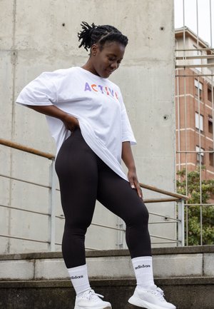 T-Shirt blanca de gran tamaño con "ACTIVE" en letras moradas y naranjas, leggings negros de canalé, zapatillas blancas y calcetines blancos de la marca Adidas.