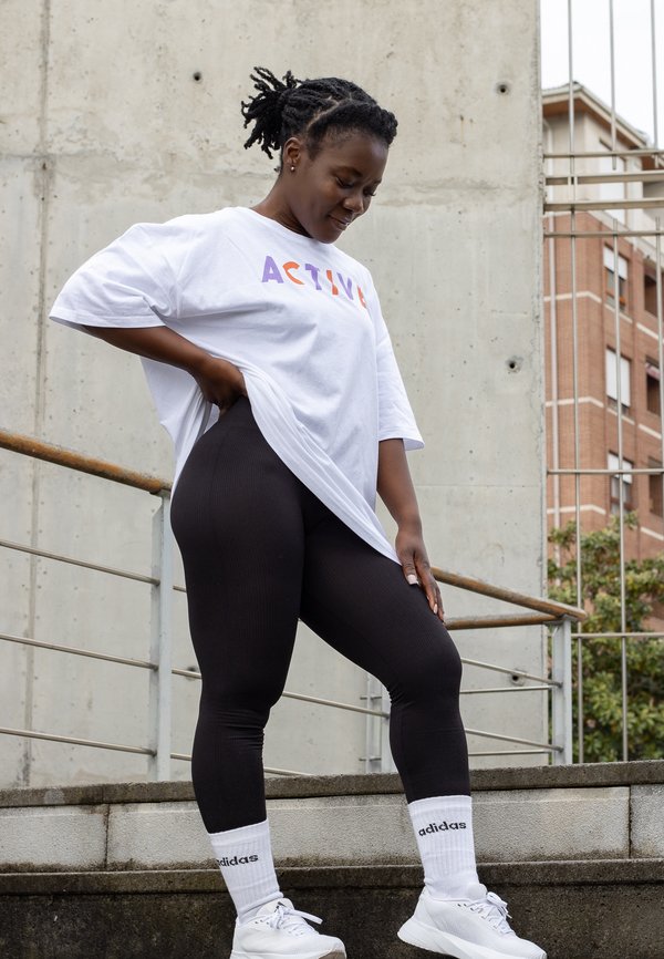 Vitt oversized t-shirt med "ACTIVE" i lila och orange bokstäver, svarta ribbade leggings, vita sneakers och vita strumpor med Adidas-logga.