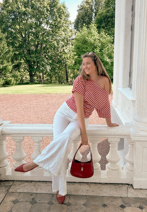 Mujer con top de un hombro a rayas rojas y blancas y pantalones blancos sentada en una barandilla de piedra blanca al aire libre, sosteniendo un bolso rojo y sonriendo.