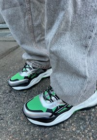 Grüne und silberne SportSneaker mit einem Mesh-Obermaterial, schwarzen Akzenten und einer klobigen weißen Sohle, kombiniert mit hellgrauen Jeans. 