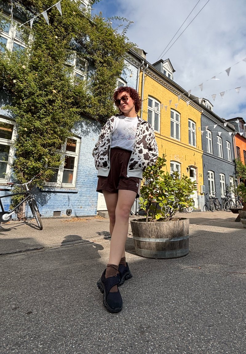 Personne portant un t-shirt blanc sous une veste bomber à motif vache noir et blanc, un short marron et des chaussures noires, se tenant dans la rue.