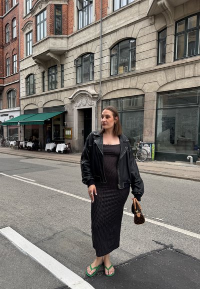 Una chaqueta bomber de cuero negra sobre un vestido midi de lunares, combinada con chanclas verdes. Sostiene un bolso pequeño en una mano. Fondo urbano de calle.