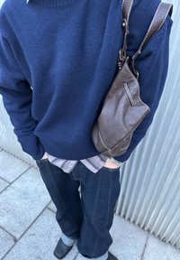 Maglione blu navy sopra una camicia a quadri, jeans in denim scuro e scarpe nere. Una borsa in pelle marrone pende dalla spalla, mettendo in mostra dettagli testurizzati.