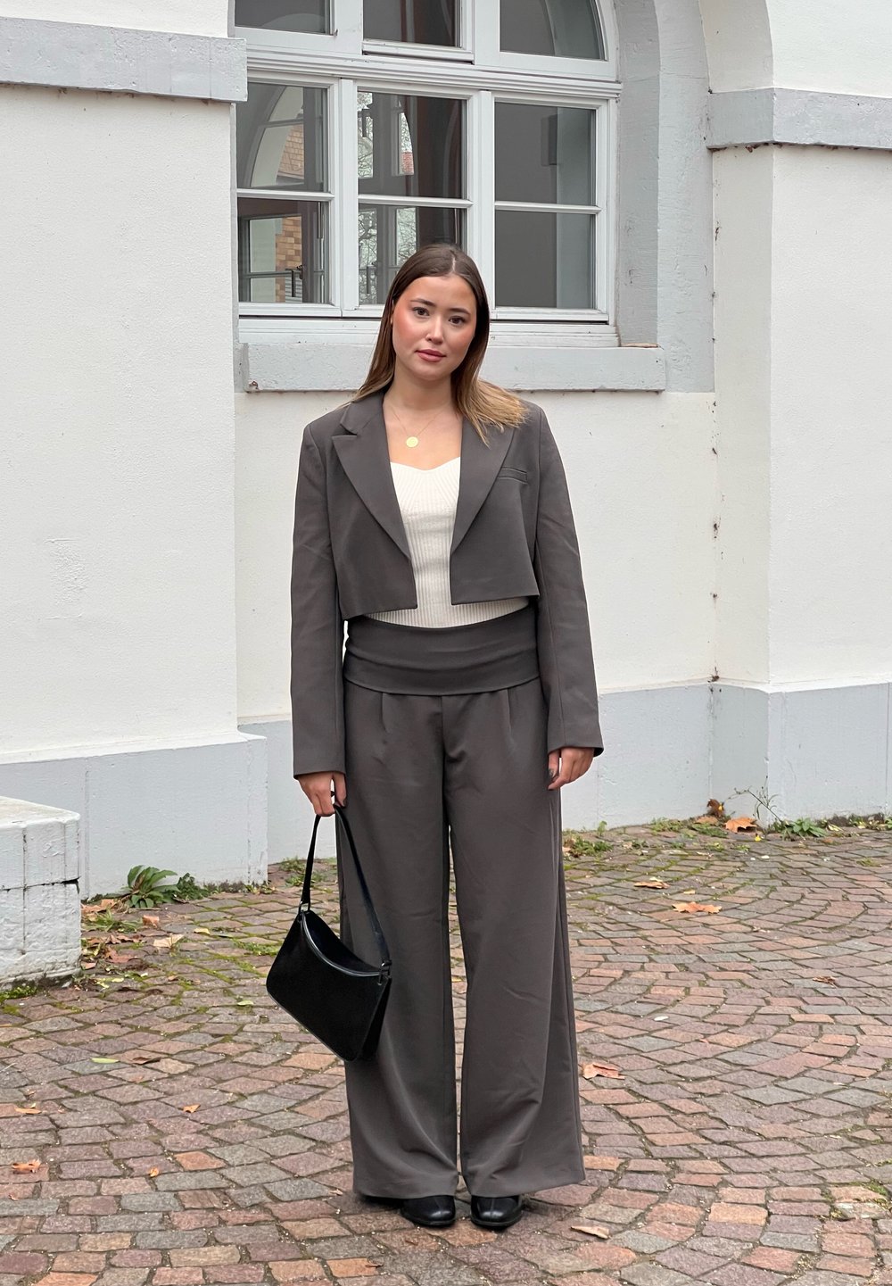 Sivina skrajna blazer in hlače širšega kroja v kombinaciji z belo prilegajočo majico. Obleko dopolnjujeta črna torba za ramo in čevlji. Teksturirana ozadje.