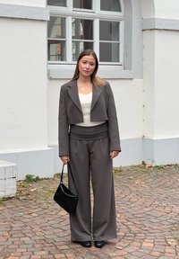 Sivina skrajna blazer in hlače širšega kroja v kombinaciji z belo prilegajočo majico. Obleko dopolnjujeta črna torba za ramo in čevlji. Teksturirana ozadje.