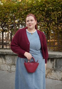 Bordowy dziergany kardigan nałożony na jasnoniebieską denimową sukienkę. Dodatki to mała czerwona skórzana torebka z uchwytem u góry i złotym zapięciem.