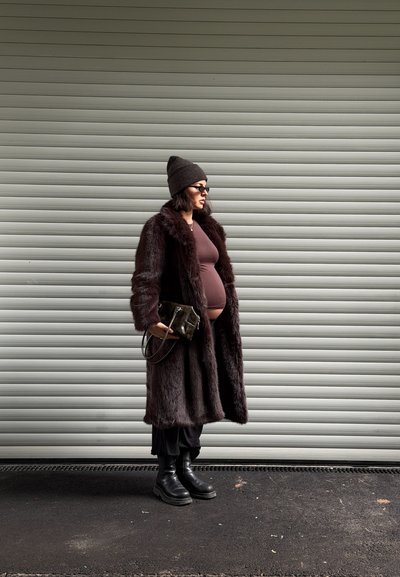 Mujer embarazada con abrigo marrón y gorro de punto sosteniendo una pequeña bolsa, de perfil frente a una puerta de metal acanalado de color gris claro.
