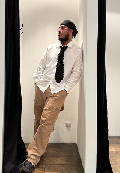 Camisa blanca, corbata negra texturizada, pantalones beiges con bolsillos, zapatos negros. Un hombre se encuentra de pie contra una pared, contrastando con la cortina oscura cercana.