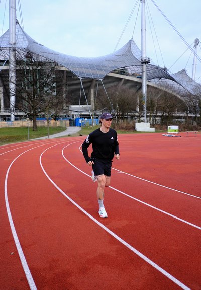 Hombre corriendo en una pista de atletismo al aire libre de color rojo, con una estructura de estadio y árboles sin hojas al fondo bajo un cielo nublado.
