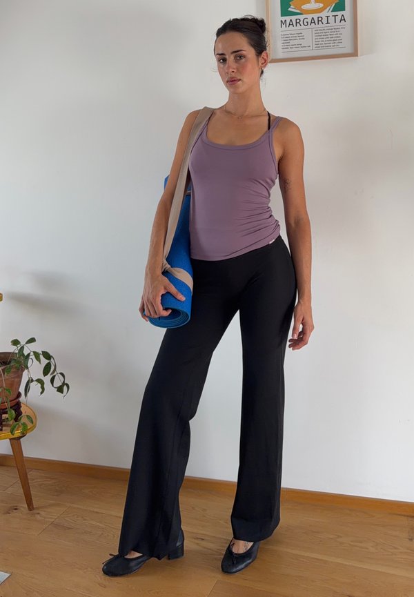 Woman standing indoors holding a blue yoga mat, wearing a purple tank top, black wide-leg pants, and black shoes.