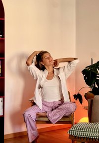 Leichte weiße Hemd über einem weißen Tanktop, kombiniert mit pastellgestreiften rosa Pyjamahosen. Sitzt im Schneidersitz auf einem Holzstuhl.