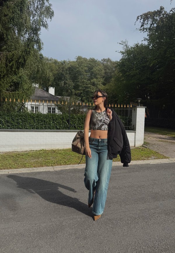 Femme marchant dans une rue portant des lunettes de soleil, un crop top, un jean à jambes larges, des chaussures marron, portant une veste et un sac à dos avec des arbres et une maison derrière.