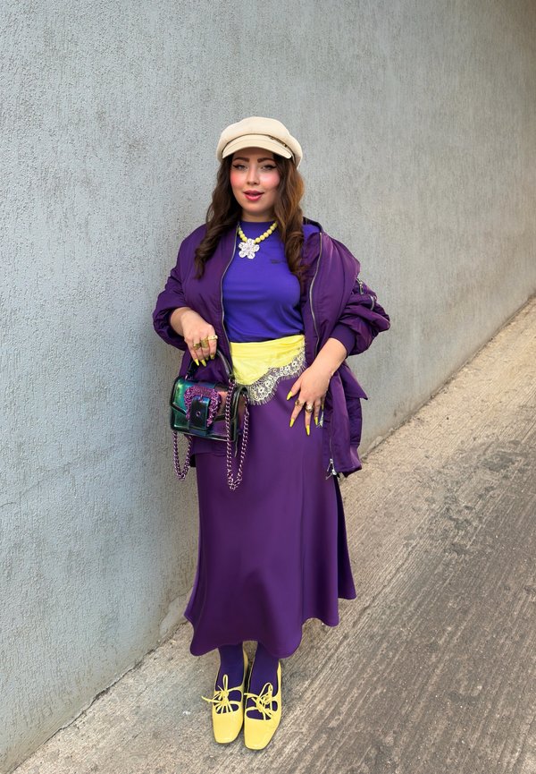 Une femme pose près d’un mur gris, vêtue d’une tenue violette, d’une casquette couleur crème, de chaussures jaunes et d’un collier, en tenant un petit sac à main irisé.