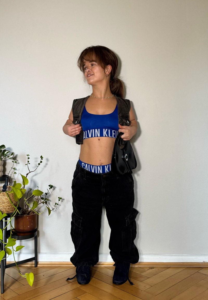 Niebieski dopasowany sportowy biustonosz i pasujący pasek, w połączeniu z luźnymi czarnymi spodniami cargo. Modelka trzyma czarną torebkę. Proste, neutralne tło.