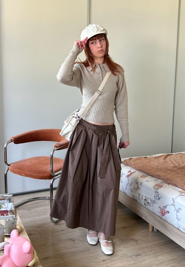 Joven mujer con suéter beige, falda larga marrón, gorra blanca y bolso bandolera, de pie en una habitación con una silla y una cama con flores.