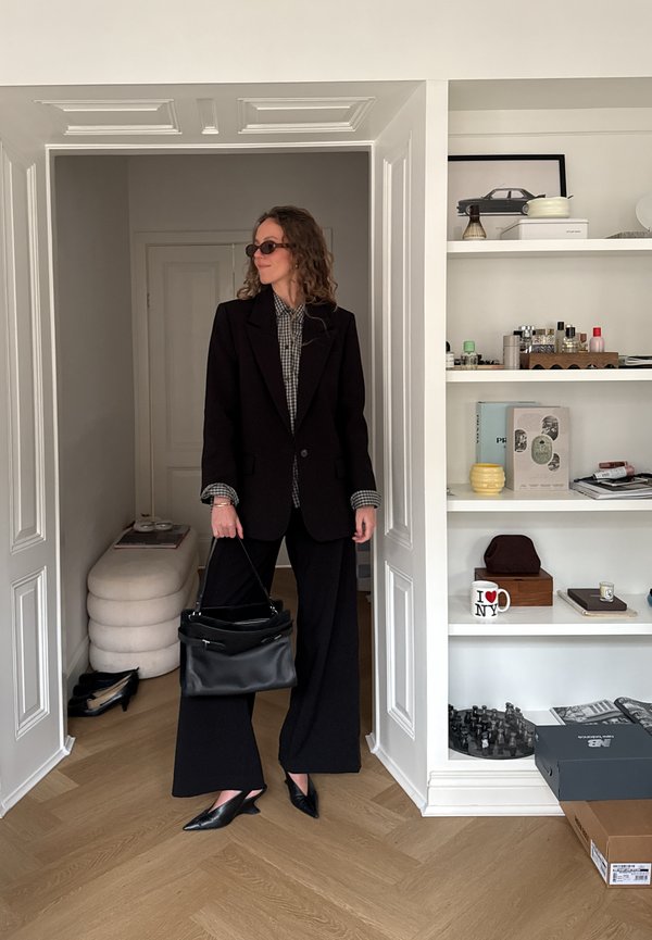 Vrouw met krullend haar, gekleed in een zwarte blazer, geruit hemd, wijde zwarte broek en zonnebril, die een zwarte handtas vasthoudt bij een deur van een moderne kamer.