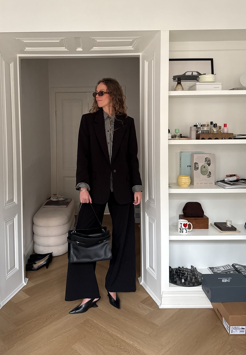 Femme aux cheveux bouclés portant un blazer noir, une chemise à carreaux, un large pantalon noir et des lunettes de soleil, tenant un sac à main noir dans l'embrasure d'une porte d'une pièce moderne.