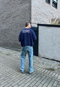 Pullover oversized blu navy con la scritta "kaotiko society" in bianco e rosa sul retro, abbinato a jeans blu chiaro e calzature casual.