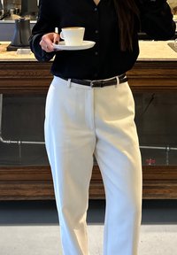 Weiße, maßgeschneiderte Hose, schwarzes Hemd mit Knöpfen, hält eine Tasse auf einer Untertasse. Verfügt über glatten Stoff, kontrastiert durch einen schwarzen Gürtel mit silberner Schnalle.